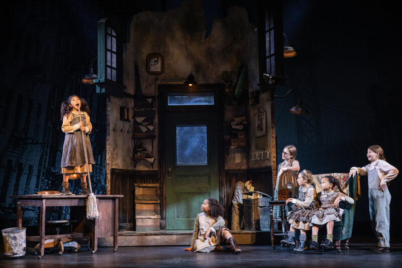 The Orphans in the North American Tour of Annie. A young girl stands on a desk singing into a mop she is using as a microphone. A group of young girls sitting on the right watch on.