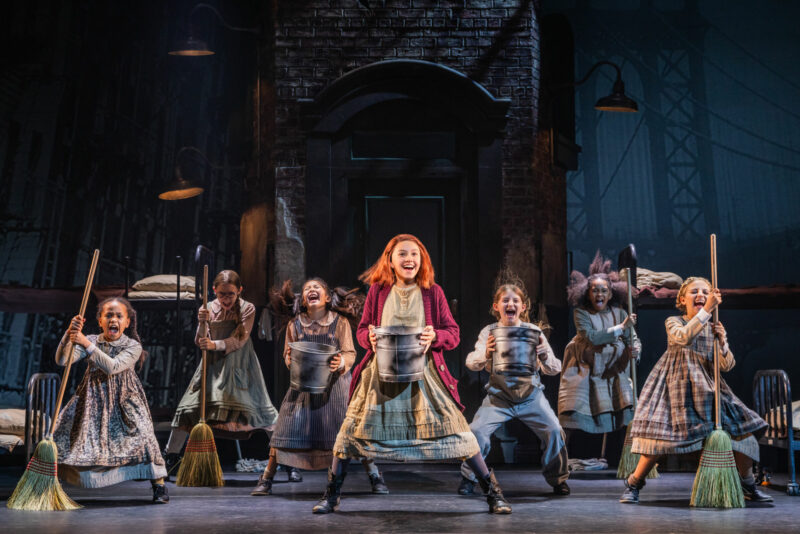 The Orphans in the North American Tour of Annie. Annie stands center holding a bucket as a group of young girls stands all around her either holding a broom or bucket.