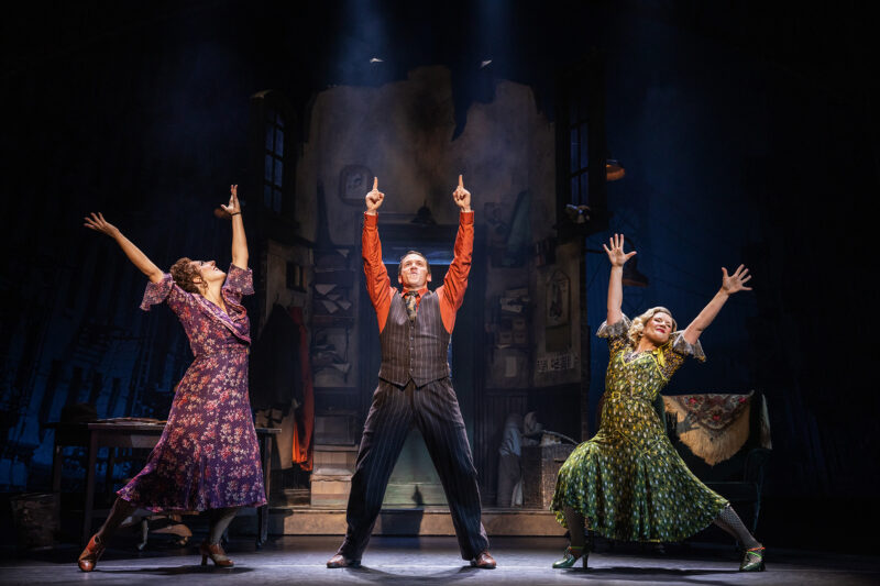 Samantha Stevens, Jeffrey T. Kelly and Stefanie Londino in the North American Tour of Annie. A man stands center between two women in the orphanage. The man points two hands in the air as the woman stretch their arms up.