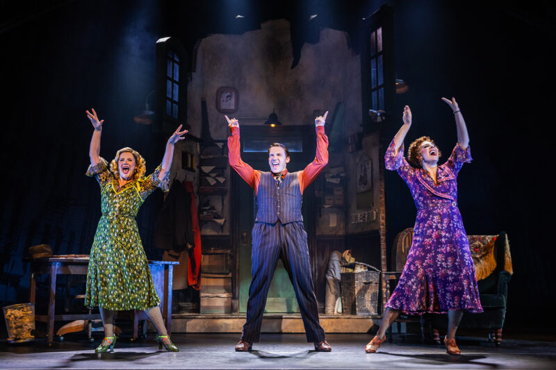 Stefanie Londino, Jeffrey T. Kelly and Samantha Stevens in the North American Tour of Annie. A man stands center between two women in the orphanage. They all seem happy as they throw hands arms in the air.