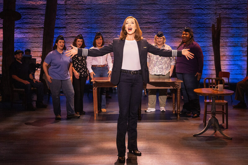 Addison Garner as ‘Beverley Bass’ and the touring cast of Come From Away. A red-haired woman wearing a pilot’s jacket stands center singing with her arms outstretched. There are five women standing behind her singing towards her, all wearing flight attendant’s hat.