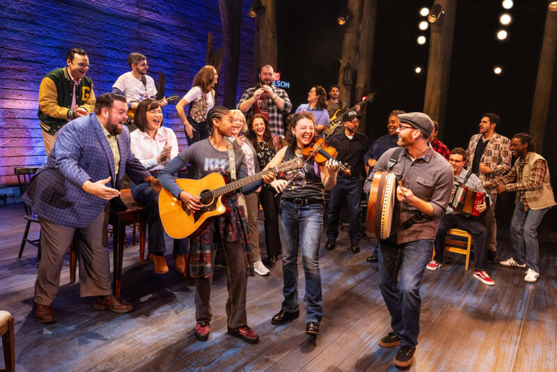 Touring Cast and Band Members of Come From Away. In the center, there a three people playing various instruments, while a large group surround them cheering them on.