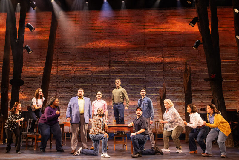 Touring Cast of Come From Away. A man in a green button-down shirt, stands atop a table, while other men kneel in front or stand beside him, all looking out towards the audience. The women are divided on either side of the table bent over in anticipation.