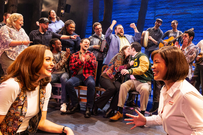 Touring Cast of Come From Away. Two women are kneeled in front of each other, laughing and smiling. There is a couple kissing behind them and a group of people cheering them on.