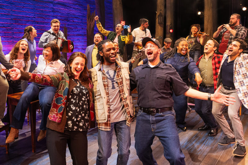 Touring Cast of Come From Away. A group of people, representing multiple identities, sing and dance together in a bar. They all are smiling with their arms around each other, and some are playing instruments.