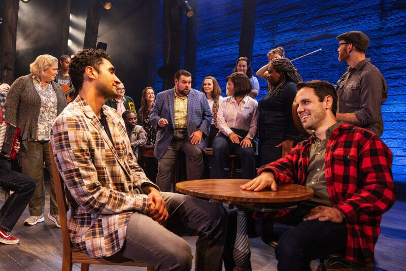 Touring Cast of Come From Away. Two men sit across from each other at a small table. A man in a suit sits behind them on a table and seems to be telling a story, as others look on. A few people are holding instruments.