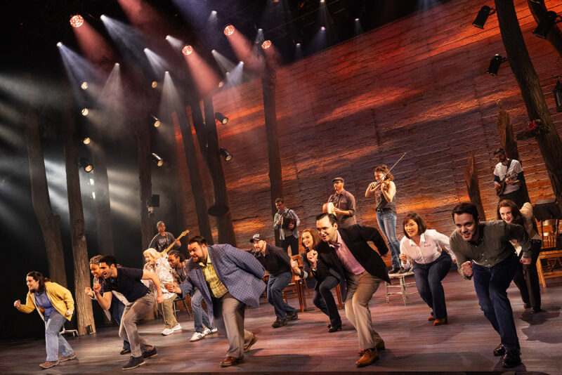 Touring Cast of Come From Away. A large group of people are all bent forward with one arm forward and one arm back, their hands in fists. People playing instruments stand on chairs in the background.