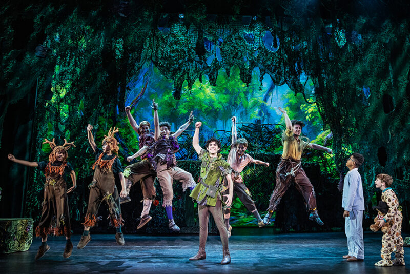 Nolan Almeida as Peter Pan and the cast of Peter Pan. Peter Pan stands center with a fist in the air and a smile on his face as The Lost Boys jump up behind him. Michael and John Darling are standing to the side looking on.